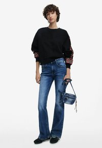 Schwarzer Sweatshirt mit gemusterten Bündchen, blaue Schlagjeans und eine kleine blaue Umhängetasche. Legere Schuhe runden den Look ab.