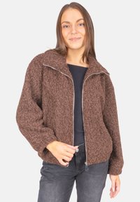 Braune, flauschige Zip-Jacke mit Stehkragen und lockerer Passform. Besitzt ein strukturiertes Material und einen silbernen Reißverschlussakzent. Wird über einem schwarzen Oberteil getragen.