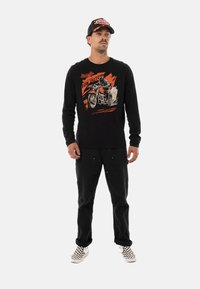 Chemise noire à manches longues avec un graphique de moto en rouge et crème. Assortie à un pantalon noir et des baskets à carreaux. Le modèle se tient sur un fond blanc.