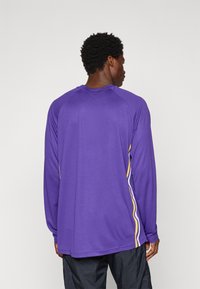 T-shirt de sport à manches longues violet en tissu texturé, avec des bandes latérales jaunes et blanches et un col rond.