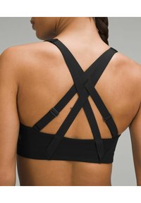 Sujeción deportiva negra con tirantes ajustables en crisscross, con una textura suave y una banda ancha para mayor soporte. Con acentos de metal notables.