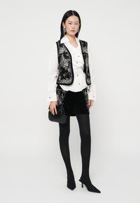 Black embroidered sleeveless vest over a white button-up shirt, paired with a black velvet mini skirt, black tights, and black heels.