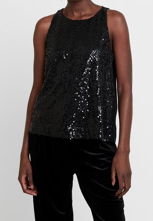 Femme portant un haut noir sans manches recouvert de sequins noirs brillants, associé à un pantalon en velours noir sur un fond uni.