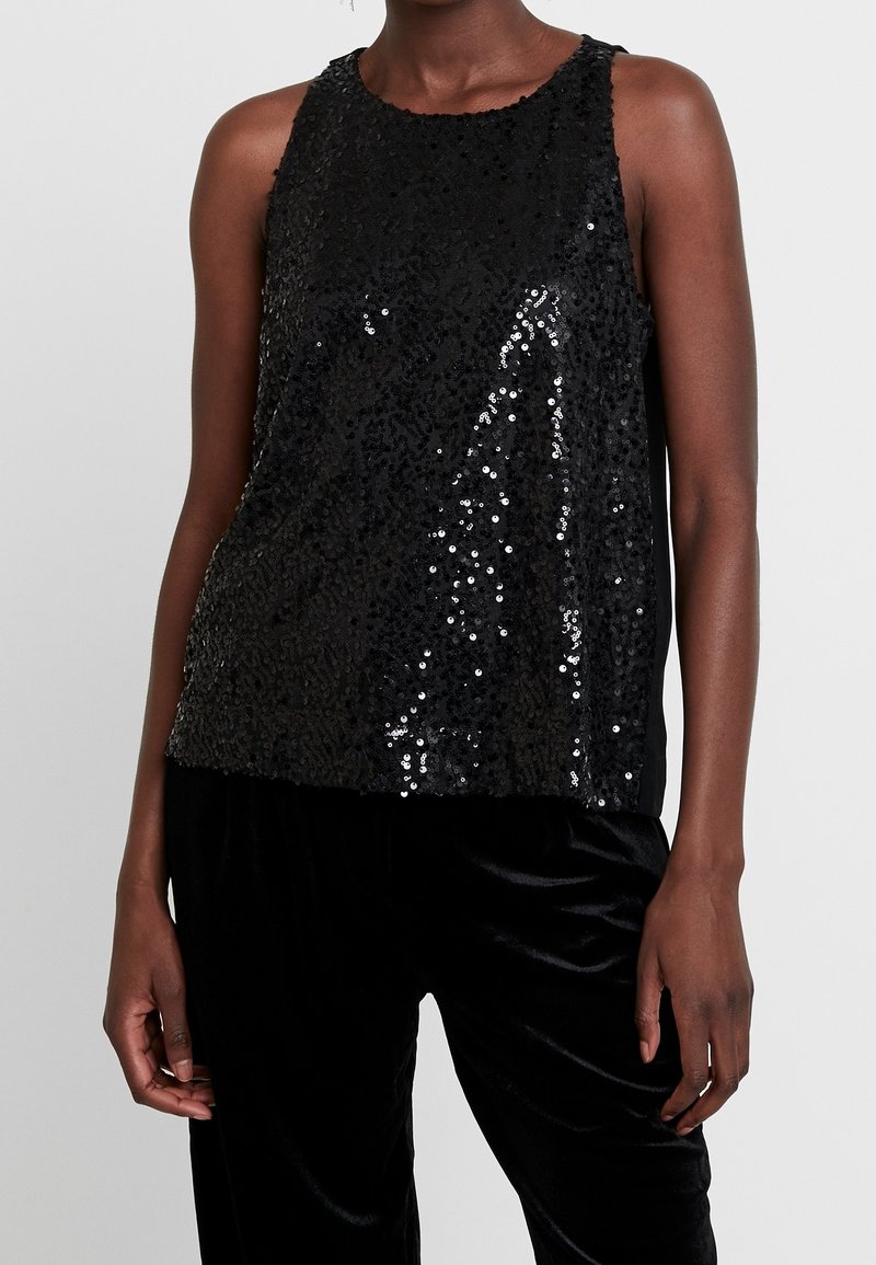 Femme portant un haut noir sans manches recouvert de sequins noirs brillants, associé à un pantalon en velours noir sur un fond uni.