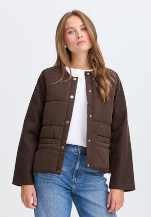 Jeune femme aux longs cheveux châtain clair porte une veste matelassée marron par-dessus une chemise blanche et un jean bleu, debout devant un fond clair uni.
