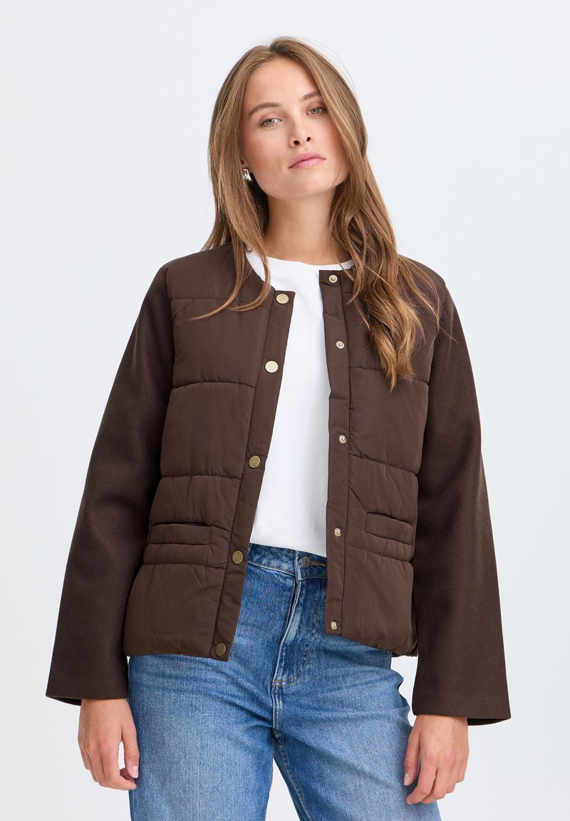 Jeune femme aux longs cheveux châtain clair porte une veste matelassée marron par-dessus une chemise blanche et un jean bleu, debout devant un fond clair uni.