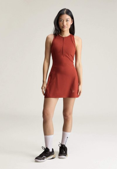 Junge Frau steht, trägt ein ärmelloses rostfarbenes Kleid, weiße Sneakersocken und schwarze Sportschuhe vor einem schlichten hellen Hintergrund.
