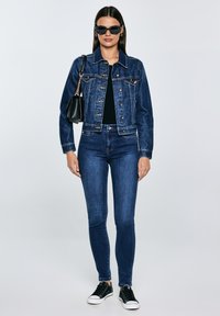 Moteris dėvi tamsiai mėlyną džinsinį švarką ir džinsus, juodą viršutinę dalį, juodus akinius nuo saulės, laiko juodą rankinę ir juodus sportbačius, stovi prieš baltą foną.