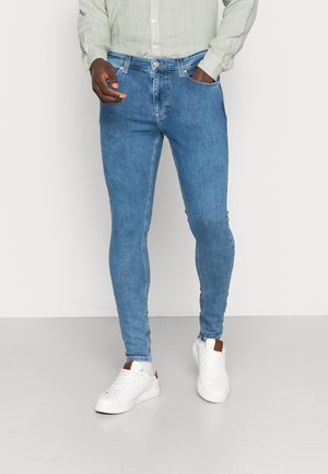 Homme portant un jean slim bleu clair, une chemise à manches longues gris clair et des baskets blanches, debout devant un fond uni.