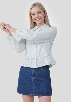 Jeune femme souriante, portant un chemisier bleu clair avec des volants et une jupe en jean bleu, étirant les bras sur les côtés devant un fond uni.