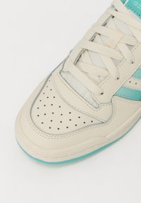 Bežīgas ādas sporta kurpes ar perforētu purngalu, kurām ir tirkīza akcenti un teksturēta zole. Šņorēm ir gaišā bežā krāsa.