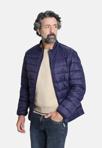 Chaqueta acolchada azul marino con diseño de acolchado horizontal, cuello alto y cierre de cremallera, llevada sobre un suéter beige y jeans azules.