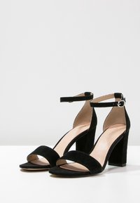 Sandales à talons hauts en cuir suédé noir, dotées de deux larges sangles avant, d'une sangle à la cheville avec une boucle, et d'un talon carré.