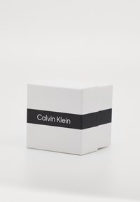 Hvit boks med et svart horisontalt bånd som har "Calvin Klein" i hvit tekst. Glatt tekstur, kvadratisk form, minimalistisk design.