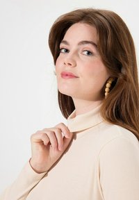 Cremfarbener Rollkragenpullover mit geripptem Muster, tailliertem Schnitt und langen Ärmeln. Das Modell justiert den Kragen mit einer Hand. Natürliches Haar und Ohrringe.