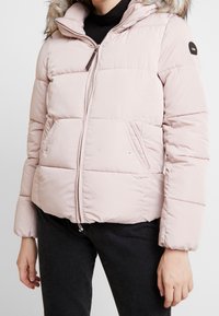 Světle růžová prošívaná bunda s kapucí lemovanou umělým kožíškem, zapínáním na zip a bočními kapsami. Prošívaný design přidává texturu a teplo.