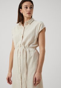 Rochie bej din in, cu mâneci scurte, închidere cu butoane și talie legată. Țesătură texturată, guler și cusături detaliate vizibile.