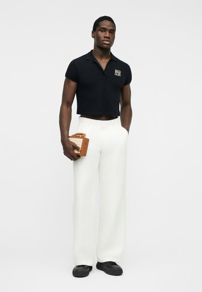 Homme portant un polo noir raccourci et un pantalon large blanc, tenant un petit sac à main marron, debout devant un fond blanc.