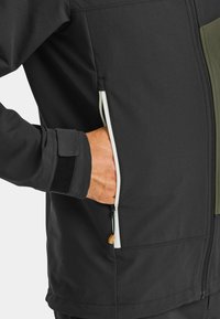 Black softshell jacket with an olive green panel, featuring a zippered pocket and velcro cuff. Durable, matte texture.