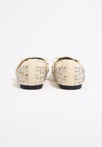 Scarpe slip-on beige con una finitura texture, decorate con borchie argentate distribuite uniformemente sul retro per un tocco di dettaglio. Suole nere.