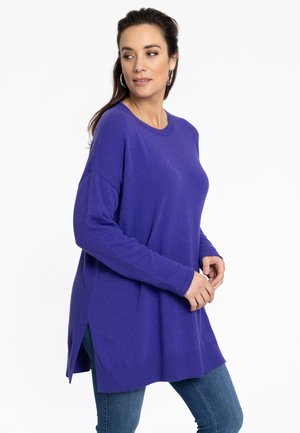 Lange mouwen, paarse trui van zacht materiaal met een ronde hals, zijsplitsingen en een relaxte pasvorm, gecombineerd met blauwe jeans.