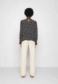 Blusa preta com padrão floral, com gola alta, detalhe nas costas em forma de buraco e mangas compridas, combinada com calças de perna larga creme de cintura alta.