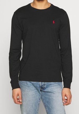 Homme portant une chemise noire à manches longues avec un petit logo rouge sur le côté gauche de la poitrine et un jean bleu clair sur un fond uni.