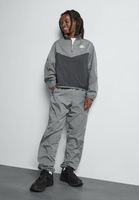 Veste de windbreaker grise Nike avec un panneau triangulaire noir, associée à un pantalon léger assorti et des chaussures de sport noires. Texture lisse, conception à fermeture éclair quart.