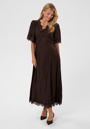Femme portant une robe midi marron foncé bordée de dentelle avec des manches courtes bouffantes, souriante et regardant vers le bas, portant des boucles d'oreilles créoles dorées et des talons noirs.
