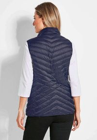 Marineblauer, gepolsterter Weste mit einem Chevron-Steppmuster, hohem Kragen und Reißverschluss. Getragen über einem weißen Oberteil, kombiniert mit schwarzen Hosen.