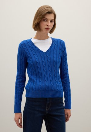 Blauer V-Ausschnitt Kabelstrickpullover mit gerippten Bündchen und Saum, kombiniert mit einem weißen Rundhalsausschnitt-T-Shirt darunter, getragen mit dunklen Jeans.