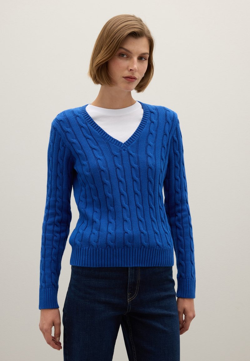 Blauwe V-hals kabelgebreide trui met geribde manchetten en onderrand, gedragen met een witte ronde hals T-shirt eronder, gecombineerd met donkere jeans.