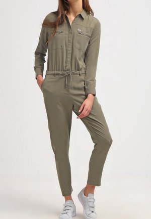 Donna che indossa una tuta a maniche lunghe verde oliva con tasche sul petto con cerniera e sneakers bianche, in piedi con le mani nelle tasche.