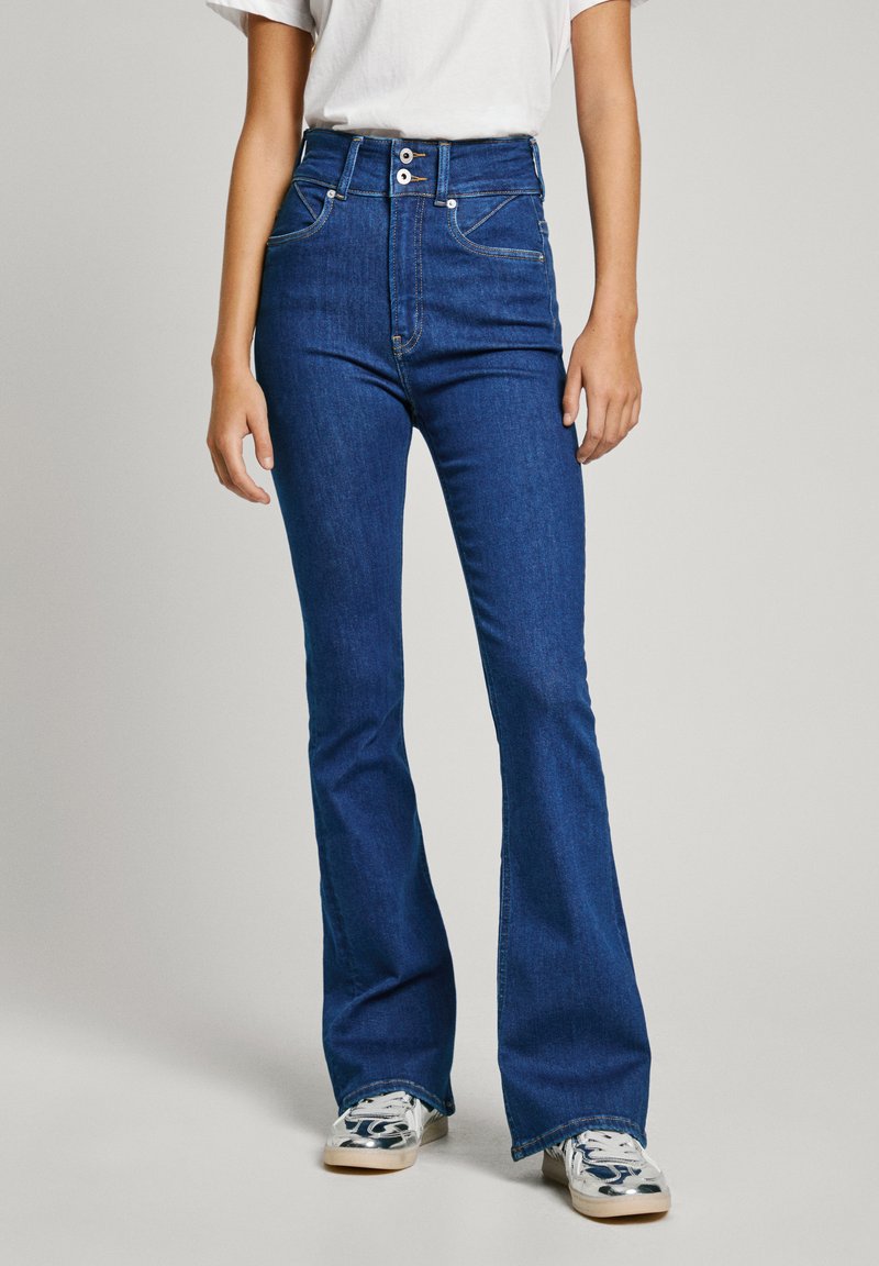 Blå högmidjade flarejeans i denim, med framfickor, tvåknappsstängning och stickade detaljer. Matchas med grå sneakers.