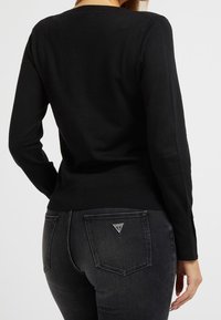 Svart långärmad tröja med ribbade kanter, i kombination med svarta jeans med en triangel-logotyp på bakfickan, slät textur.