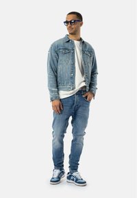 Chaqueta de mezclilla con detalles desgastados, camiseta blanca, jeans azul claro y zapatillas azules con cordones blancos. Gafas rectangulares azules.