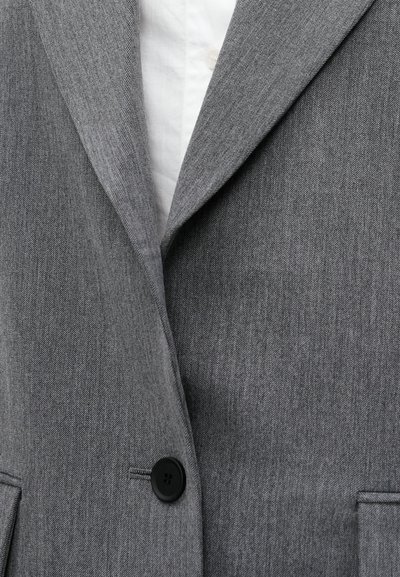 Blazer gris ajusté en tissu texturé. Comprend des revers crantés, une fermeture à un seul bouton noir et des poches avant. Une chemise blanche bien repassée est visible en dessous.