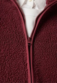 Bordeauxrode fleecejas met rits en structuurstof, voorzien van een rits aan de voorkant en een witte overhemdkraag die eronder zichtbaar is.