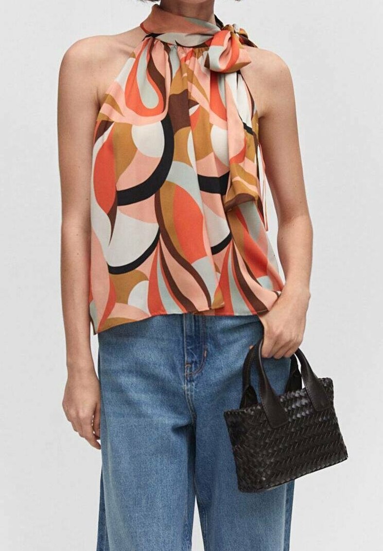 Femme portant un haut dos nu abstrait multicolore avec un nœud au cou, un jean bleu, et tenant un petit sac à main noir tressé dans la main droite.