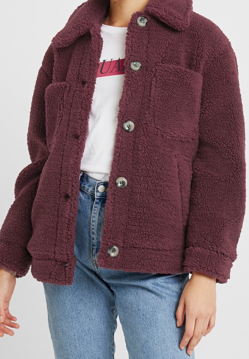 Persona vistiendo una chaqueta burgundy de botón esponjosa sobre una camiseta gráfica blanca y jeans azules, de pie contra un fondo liso.