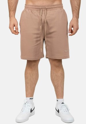 Mann trägt beige Shorts mit Kordelzug, weiße Puma-Socken und weiße Nike-Sneaker und steht vor einem einfarbigen Hintergrund.