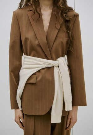 Mujer con blazer marrón de rayas finas y suéter blanco atado a la cintura, de pie frente a un fondo claro y sencillo.