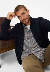 Marineblauer Grobstrick-Cardigan mit umklappbarem Kragen, kombiniert mit einem karierten Hemd in Braun und Weiß sowie beigen Hosen. Lässige Umgebung.