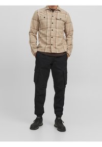 Beige rutigt knäppt skjorta med två bröstfickor över en brun t-shirt, tillsammans med svarta cargo-byxor och svarta träningsskor.