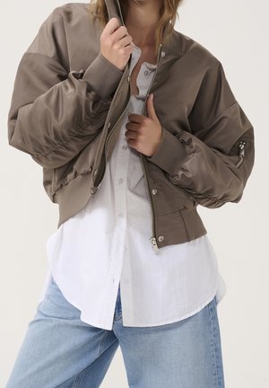 Personne portant une veste bomber taupe sur une chemise blanche boutonnée et un jean bleu clair, les mains ajustant la fermeture éclair de la veste.
