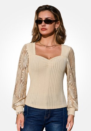 Kobieta w kremowym prążkowanym topie z długimi rękawami z kwiatowej koronki, ciemnych okularach przeciwsłonecznych, złotym naszyjnikiem i niebieskich dżinsach na białym tle.