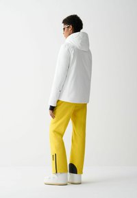 Jachetă albă impermeabilă cu glugă, asortată cu pantaloni de schi galbeni strălucitori cu un accent negru și logo, purtată cu cizme albe.