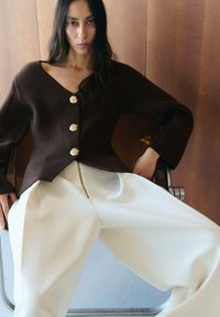 Frau mit langen dunklen Haaren, die einen dunkelbraunen, geknöpften Pullover und weite weiße Hosen trägt, sitzt auf einem Metallstuhl vor einer Holzwand.