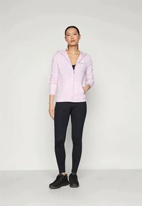 Mujer de pie con una sudadera ligera de color rosa claro con cremallera, leggings negros y zapatillas negras contra un fondo claro y liso.