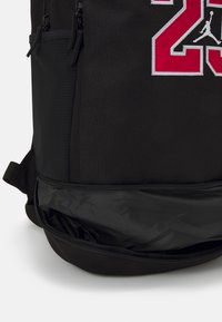 Zaino nero con tessuto strutturato, tasche con zip e ricamo rosso del "23" e logo Jordan sul fronte. Comparto aggiuntivo nella parte inferiore.